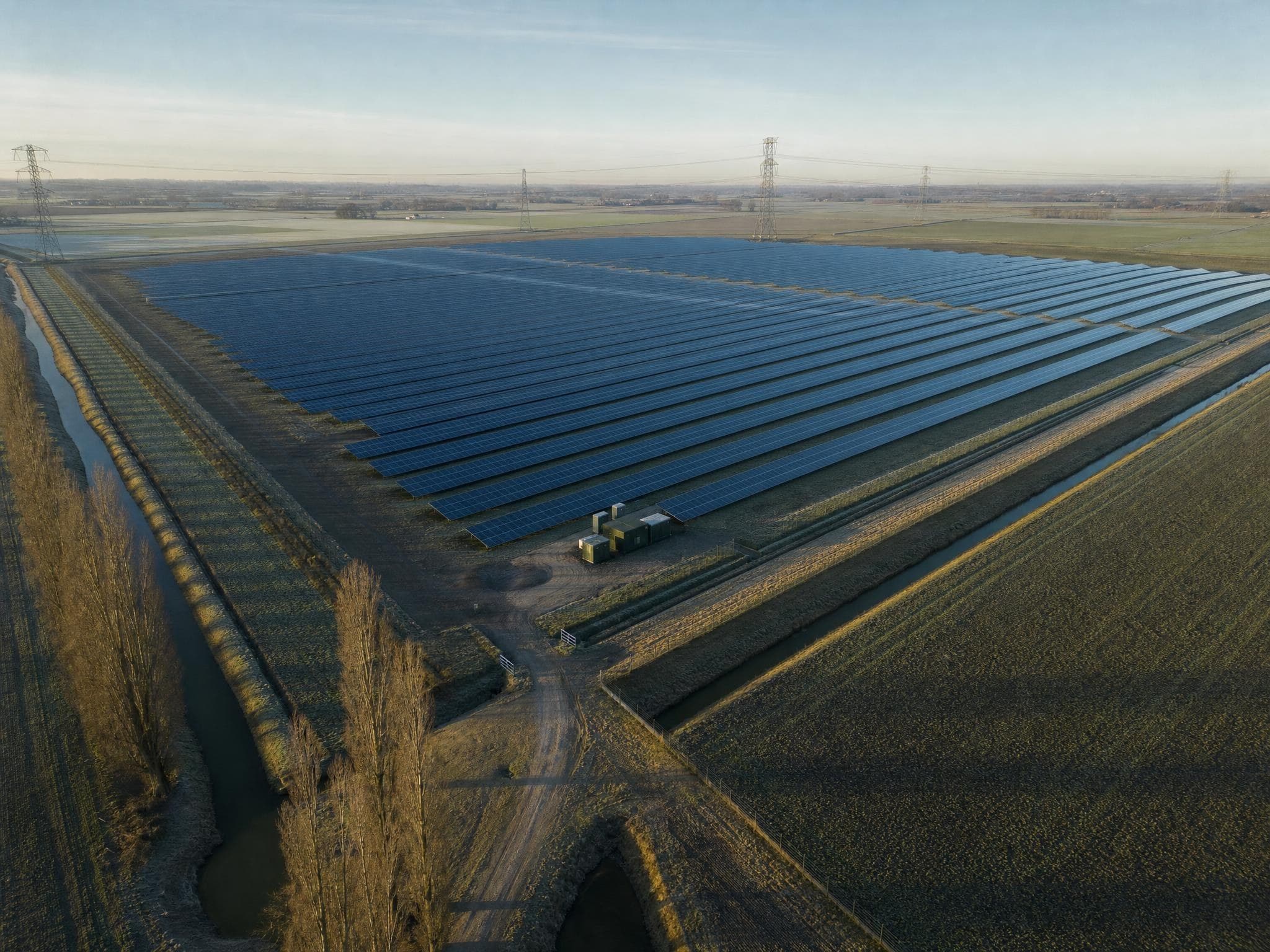 Aerial view of a solar park in a flat landscape, with a high-voltage line on the horizon