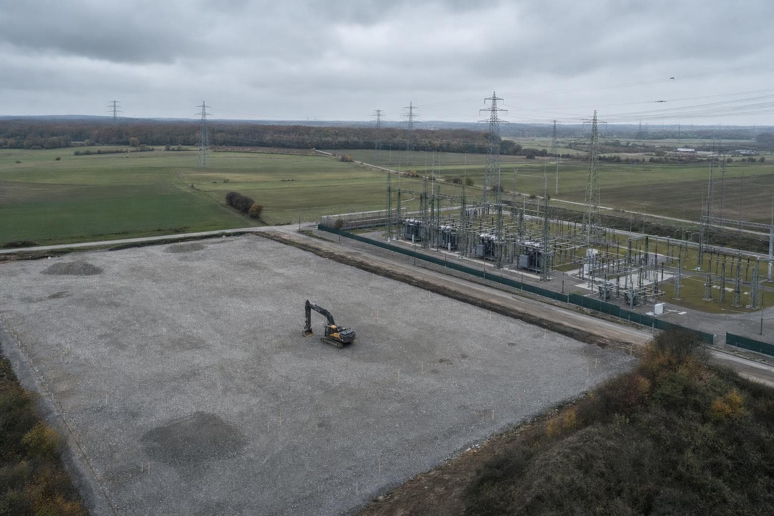 Luftaufnahme einer geschotterten Freifläche mit Vermessungsstäben und einem Bagger, direkt daneben ein Umspannwerk mit Freileitungen